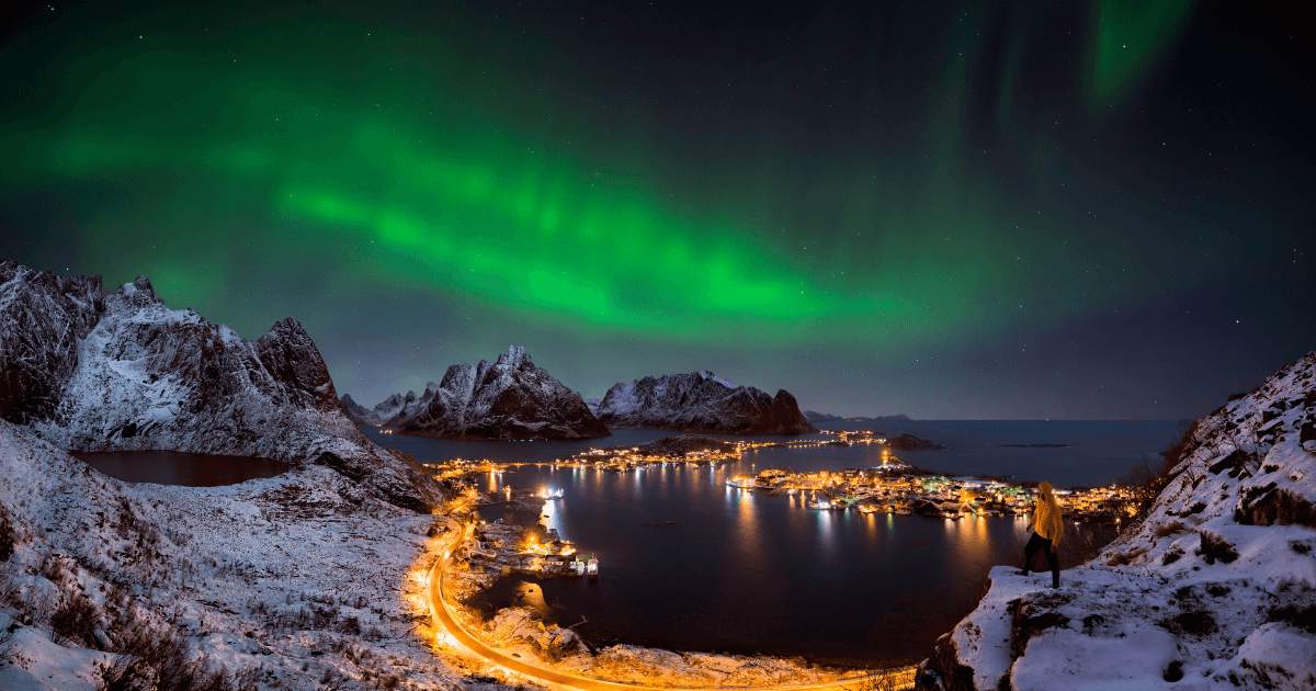 夜晚極光照亮雪山與海灣小鎮，一名旅人站在高處欣賞北極光與冬季雪景。